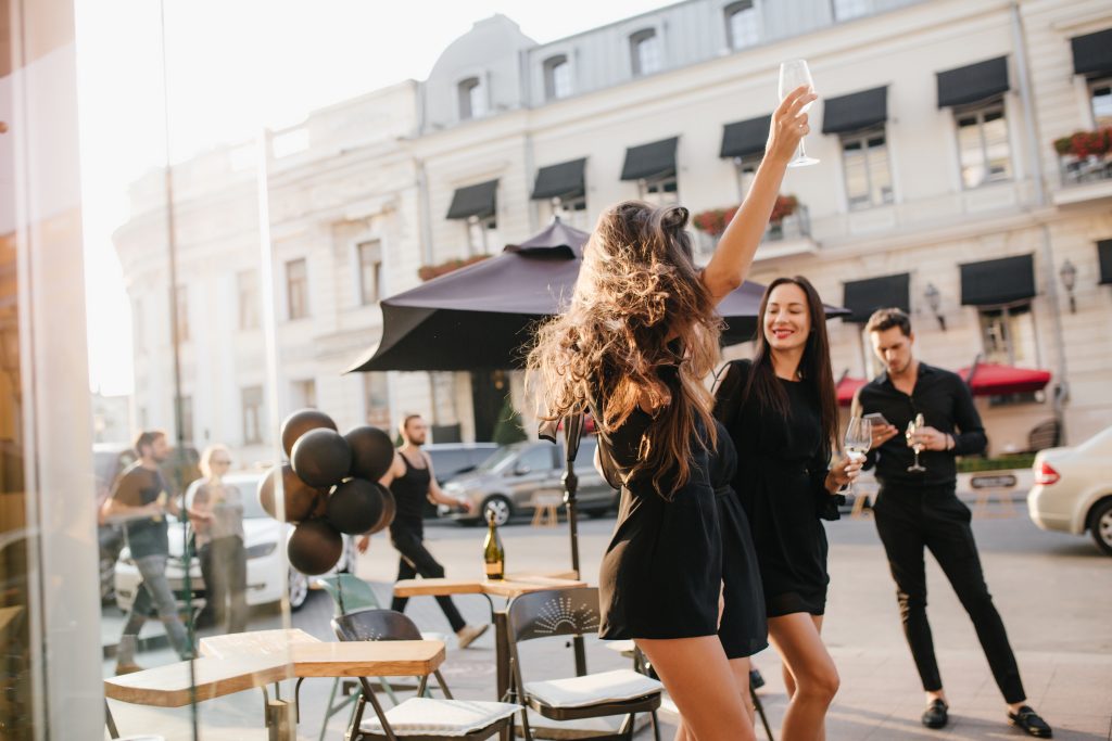 Outdoor portrait of dancing tanned female model with long hair waving. Slim girl with romantic hairstyle fooling around in street cafe and looking at young man in black attire holding glass of wine..